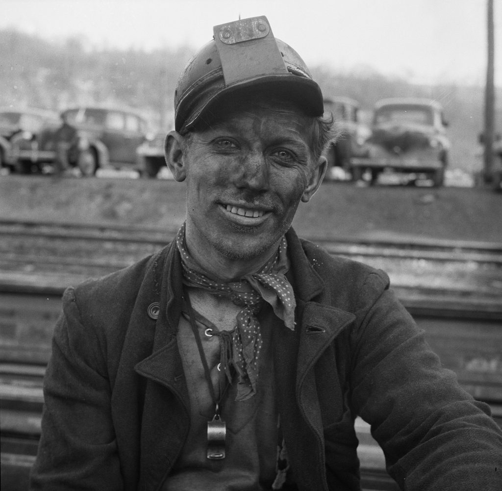 Black-and-white portrait of a coal miner, coal dust on his face, smiling at the camera, helmet lamp bracket fitted.